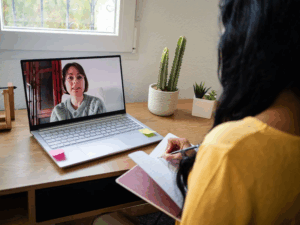 Woman attending virtual therapy session on laptop and taking notes, representing virtual IOP and personalized mental health care at Rooted Stillness in Newport Beach